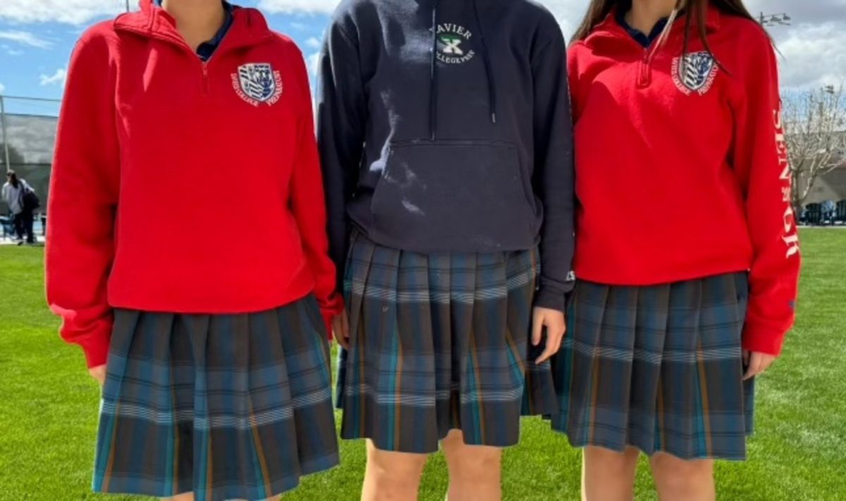 Xavier girls pose in their plaid in the grass by the tennis courts. The red senior sweatshirts are featured on the left and right, with blue polos underneath and blue plaid skirts on the bottom. Uniforms are an important part of Xavier as they make all students equal and they instill discipline. 